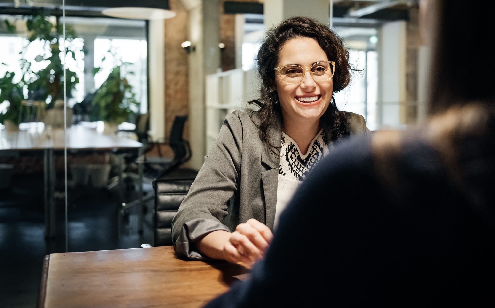 A woman with dark hair and glasses is sitting at a table during a business meeting. She seems happy and is smiling.