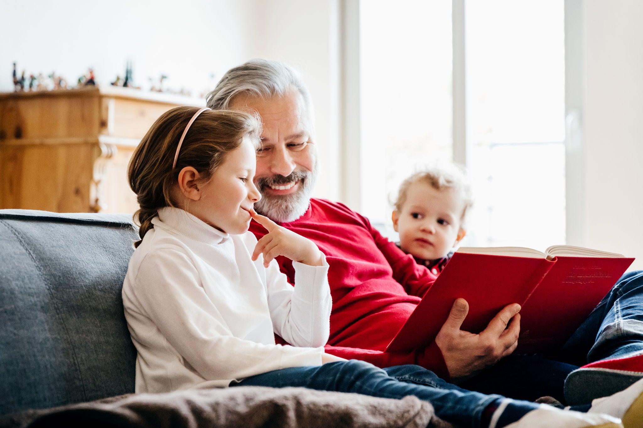 A grandpa sitting on the sofa reading a festive book to his two young grandchildren.