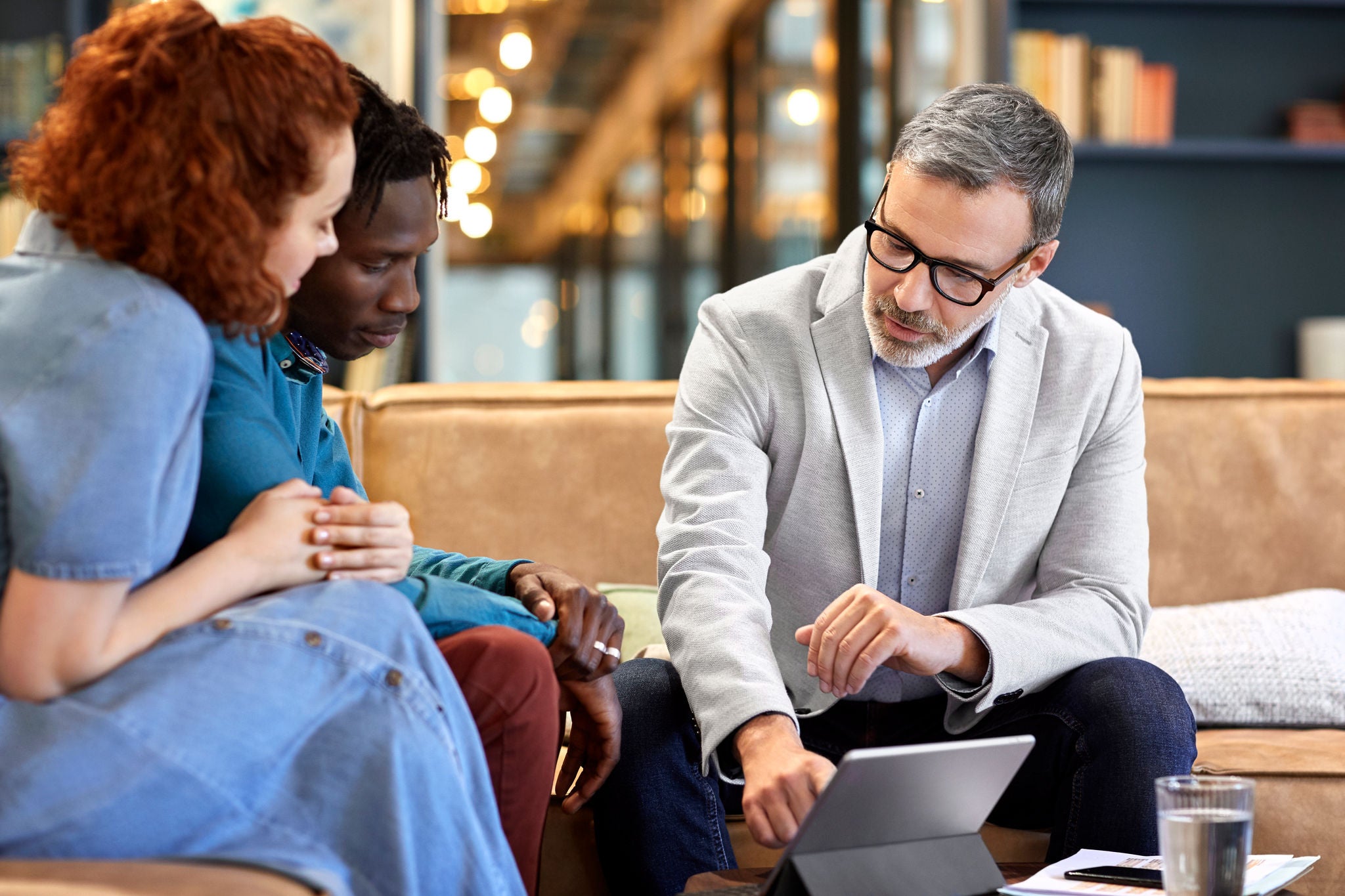 Mature financial advisor planning with young multi-ethnic couple at office. Businessman is discussing over digital tablet with clients at workplace. They are sitting in meeting.